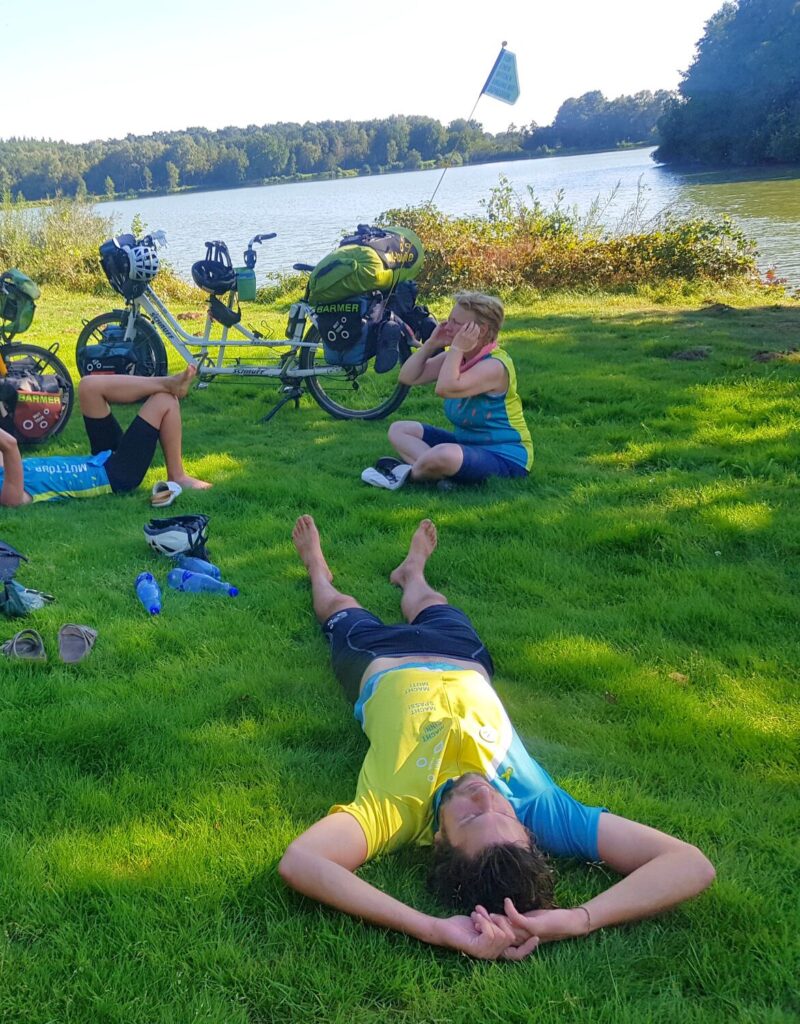 Drei Menschen sitzen und liegen auf einer Wiese am Fluss zwischen den Tandems und machen Pause