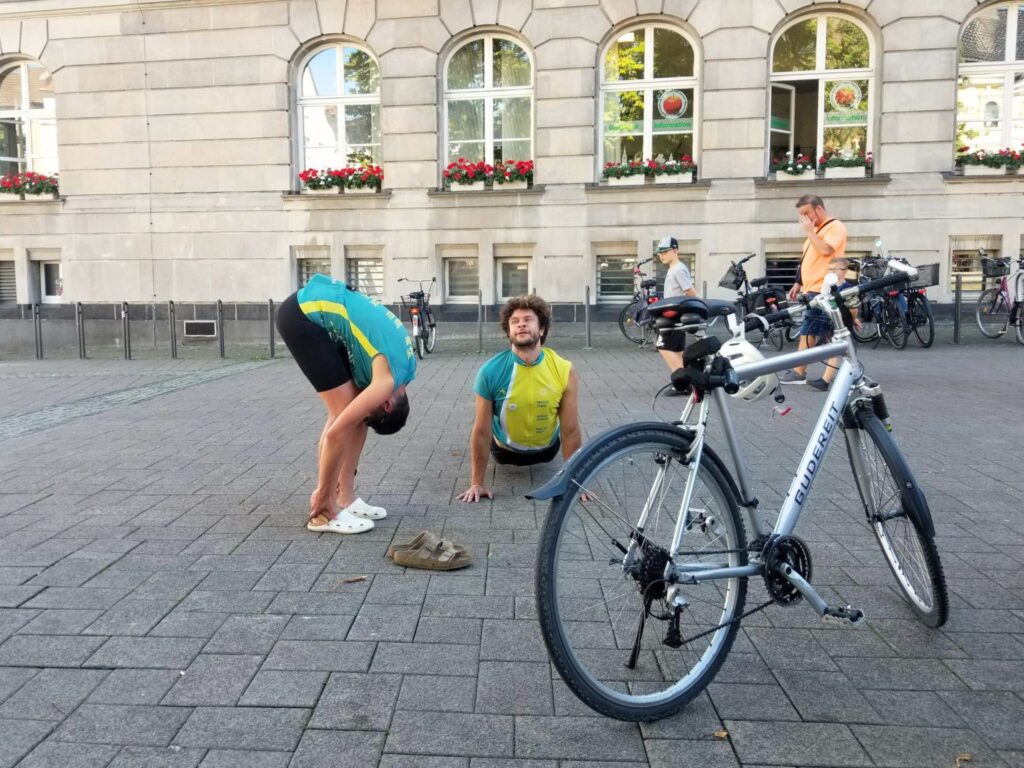 Zwei Menschen mit MUT-TOUR-Trikots machen Aufwärmübungen, neben ihnen steht ein Fahrrad